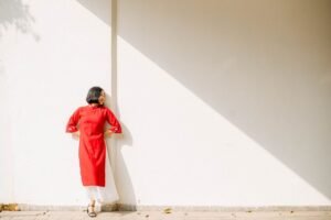 A woman in a red dress stands against a wall, bathed in bright sunlight, creating a striking shadow.