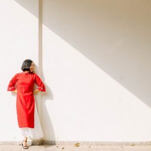 A woman in a red dress stands against a wall, bathed in bright sunlight, creating a striking shadow.